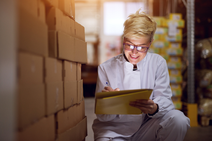 A young happy female worker is checking a list next to a stack of corrugated boxes, demonstrating the importance of good product packaging.